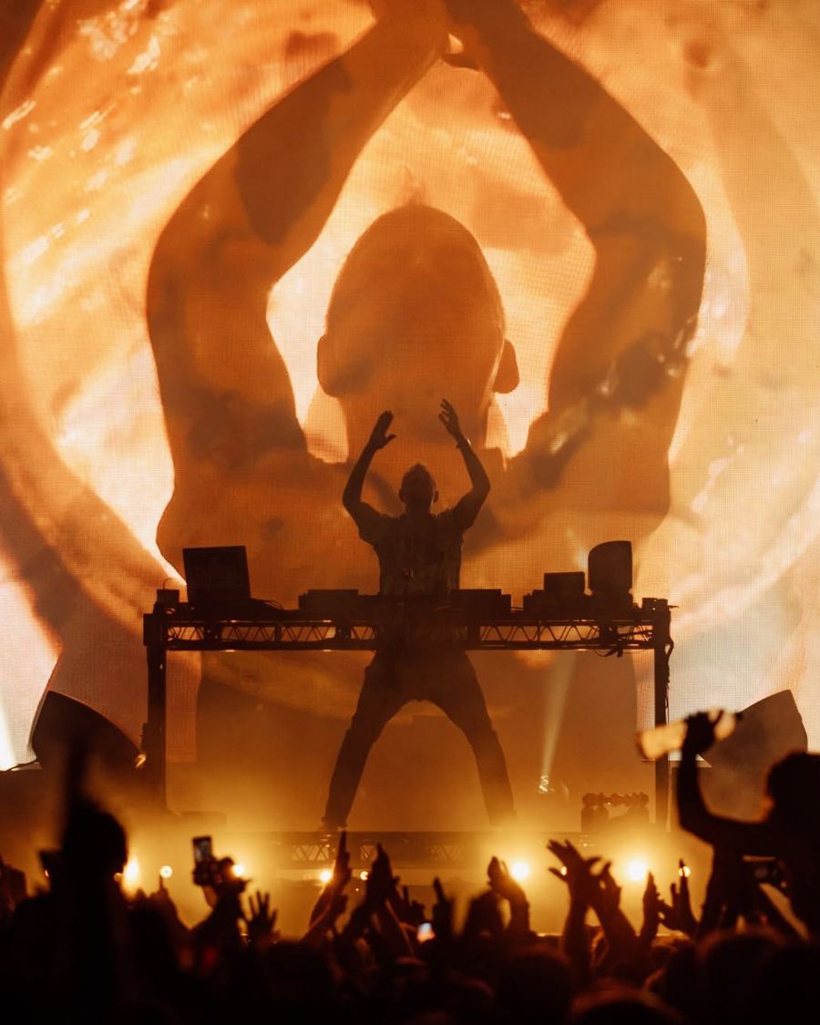 Fatboy Slim silhouetted at the decks against a giant orange screen, crowd hands raised