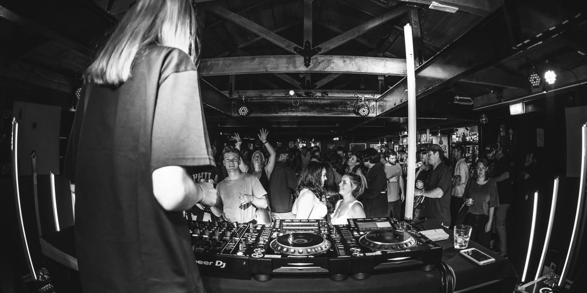 Black and white photo from behind a DJ looking out at a busy dance floor under timber beams